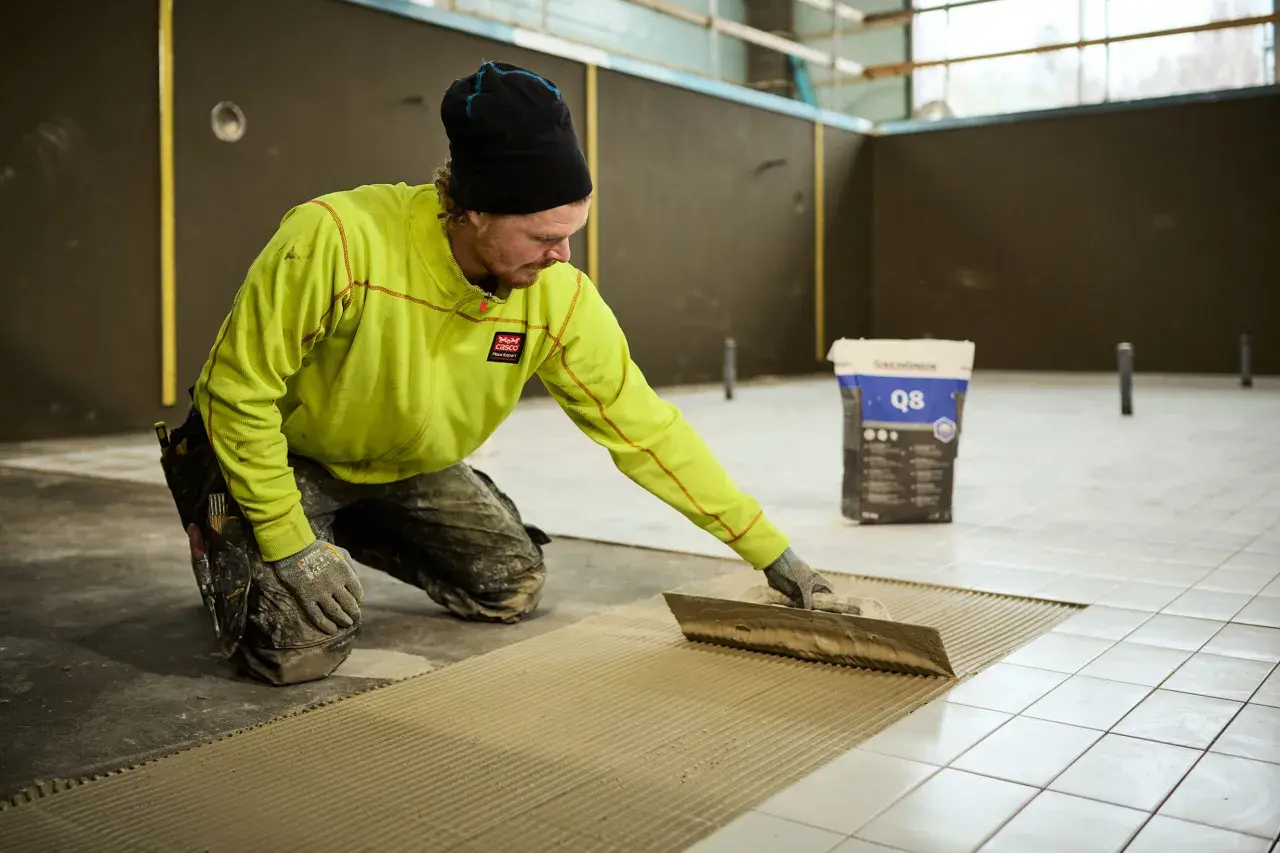 Man i gul jacka och mössa sprider ut fix med en tandspackel för att byta golv. En säck med fix står i bakgrunden.