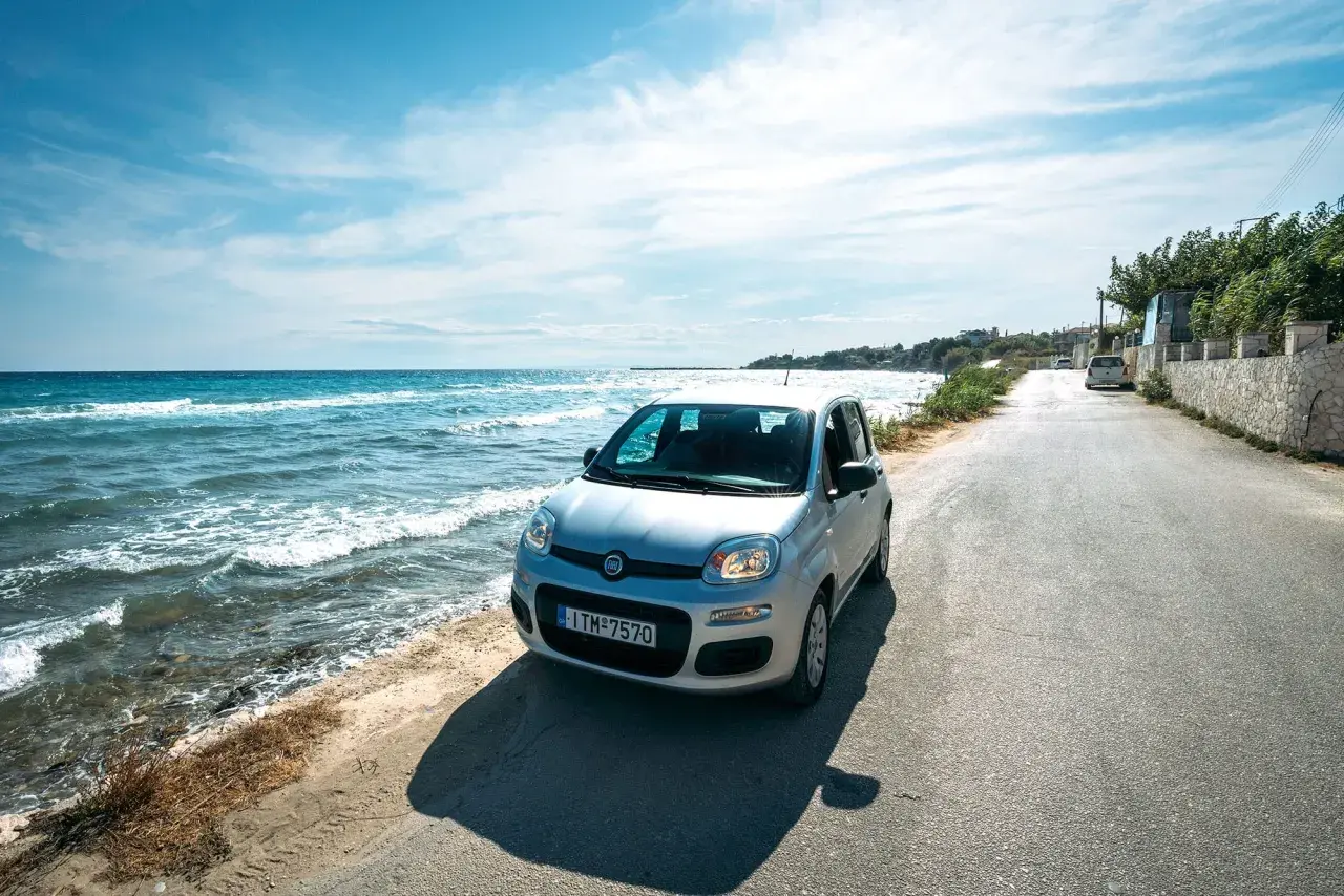 A silver Fiat Panda parked on a coastal road in Greece. The turquoise sea laps the shore beside the car, suggesting this is the perfect way to explore.
