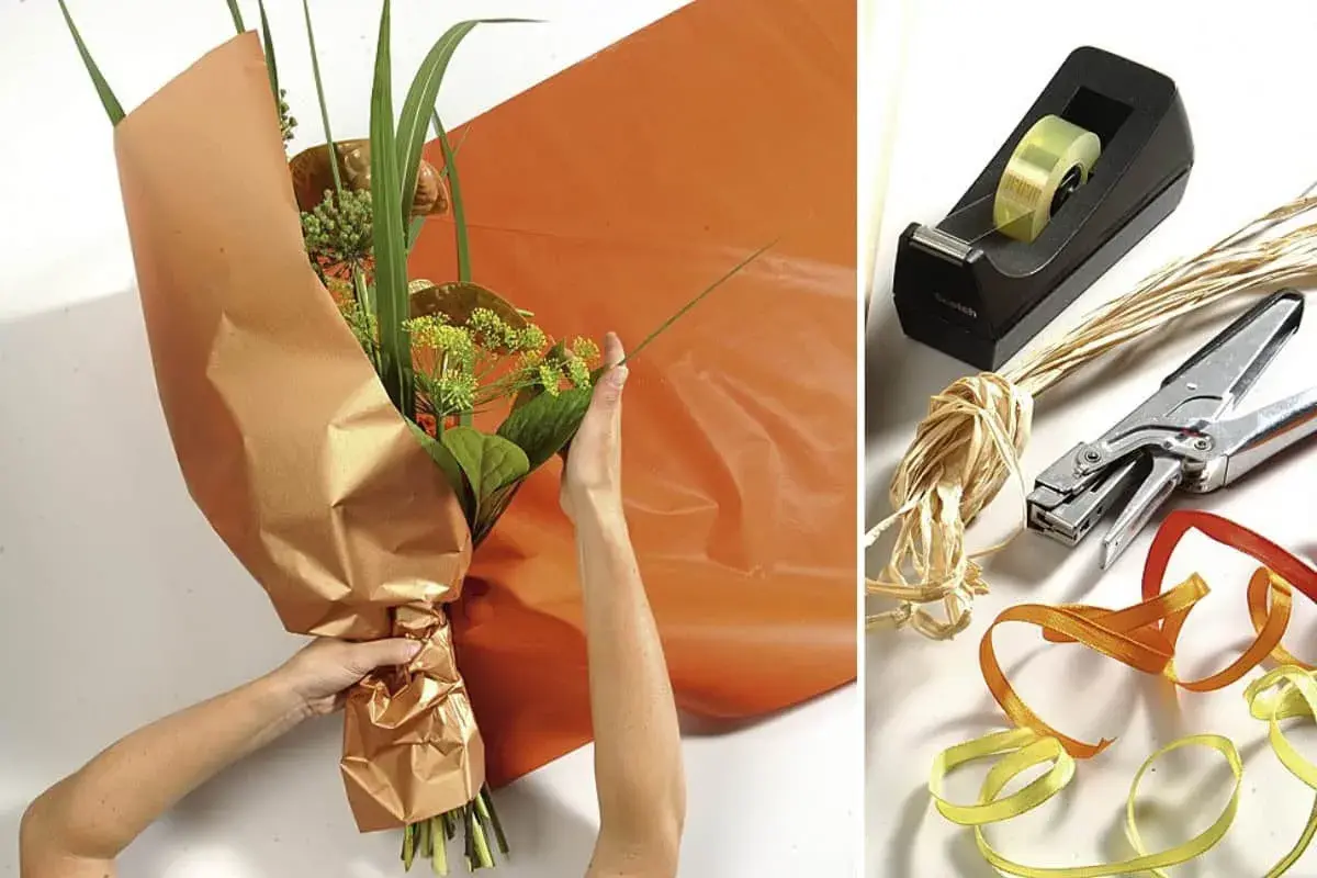Anleitung: Blumen mit Packpapier verpacken. Hände arrangieren den Strauß, während orangefarbenes Papier bereitliegt. Werkzeuge wie Klebeband, Schere und Bänder sind daneben zu sehen.