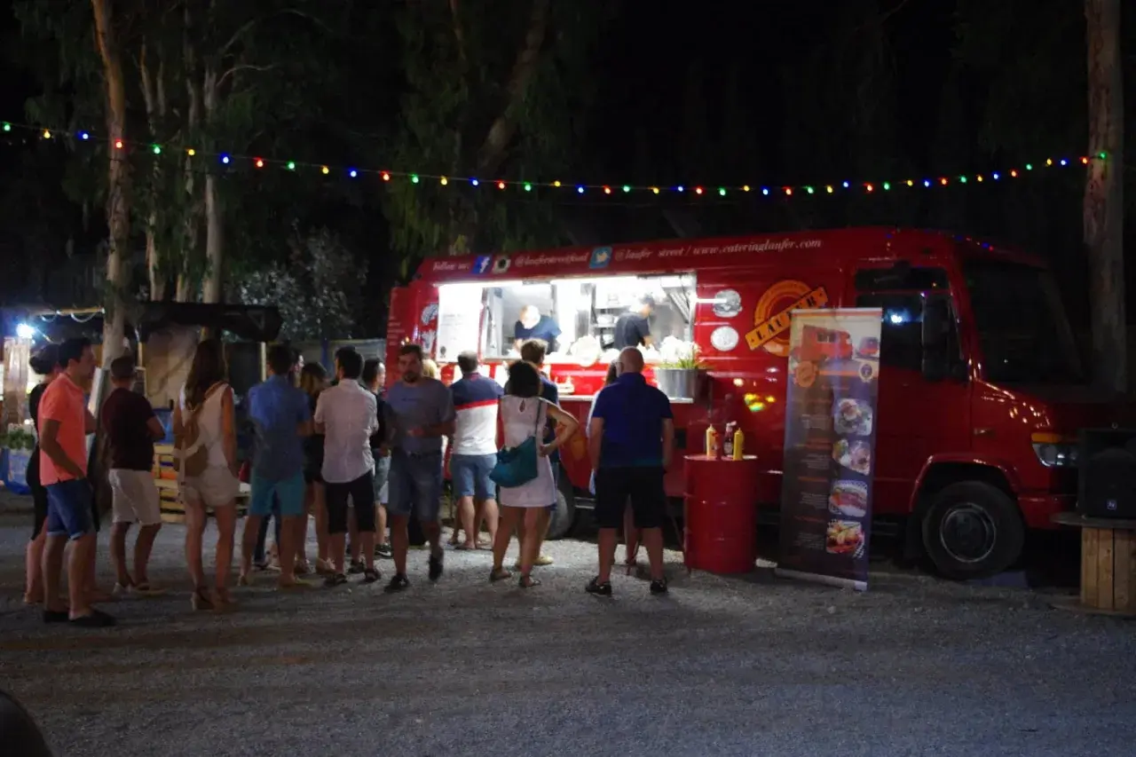 Gente disfrutando en un food trucks festival bajo luces de colores. Un camión rojo sirve comida mientras clientes esperan.