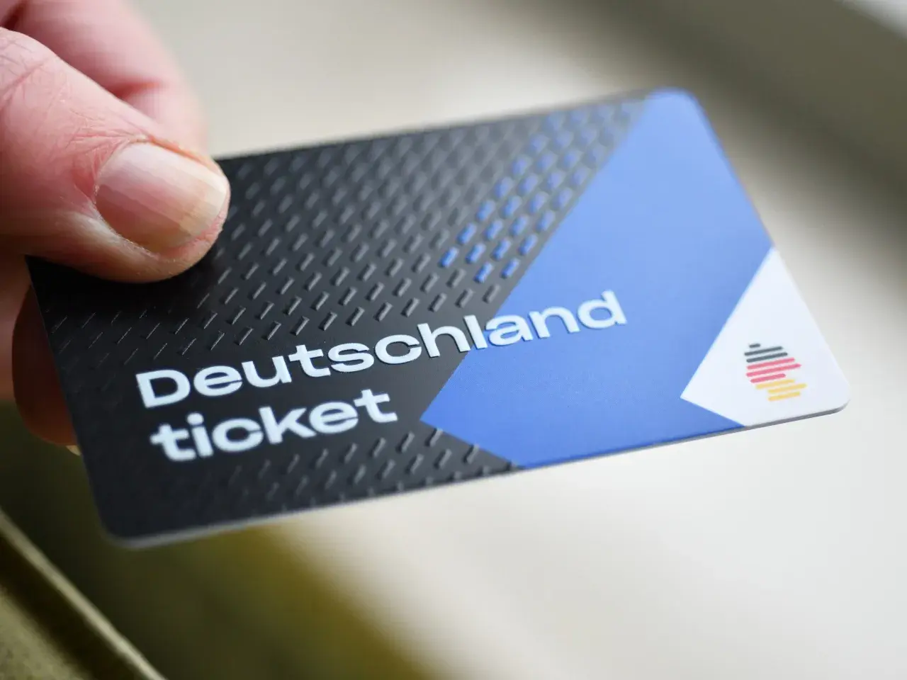 Ein Daumen hält ein Deutschlandticket. Das Ticket hat ein schwarzes, strukturiertes Design mit einem blauen und weißen Bereich.