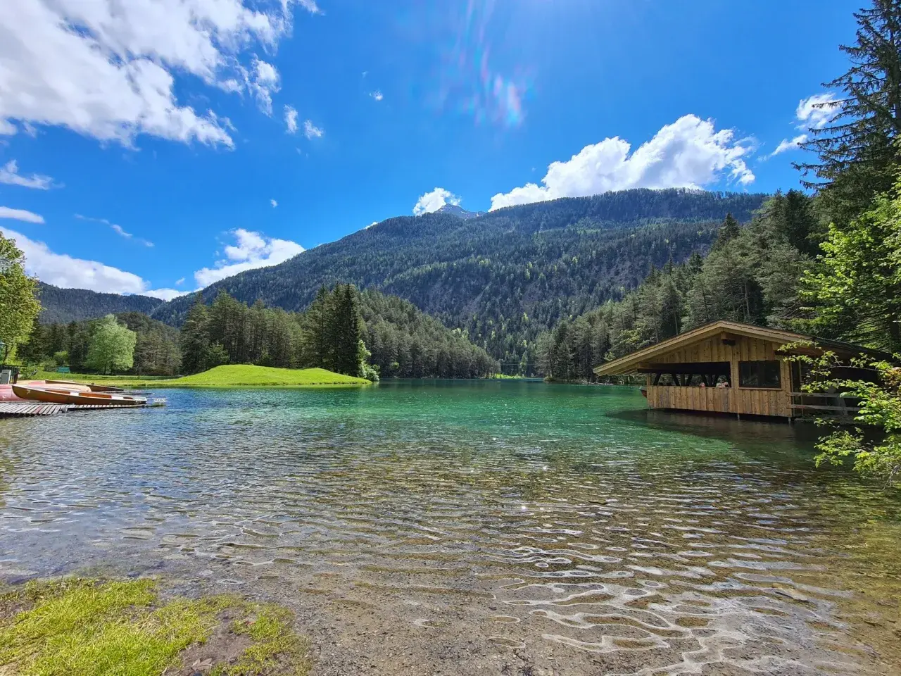 Ein idyllischer See mit türkisfarbenem Wasser, umgeben von grünen Hügeln und Wäldern. Ein Holzsteg mit Booten und ein kleines Holzhaus am Ufer laden zum Verweilen ein. Perfekt für einen blindsee trail.