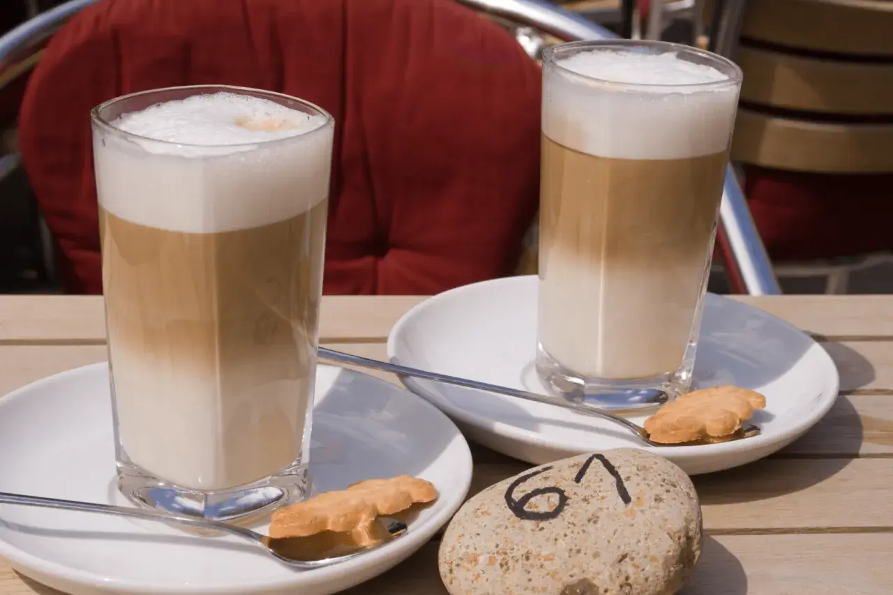 Dwa latte macchiato, czyli kawy z warstwami mleka i pianki, na stoliku. Obok ciasteczka i kamień z numerem 61.