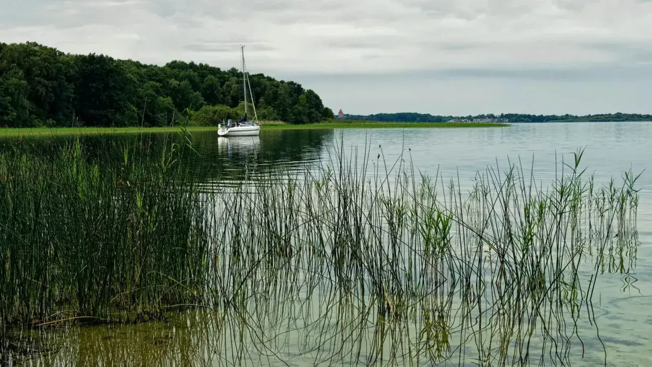 Żaglówka na spokojnym jeziorze, otoczona zielenią. Idealne miejsce na tygodniowy rejs po Mazurach.