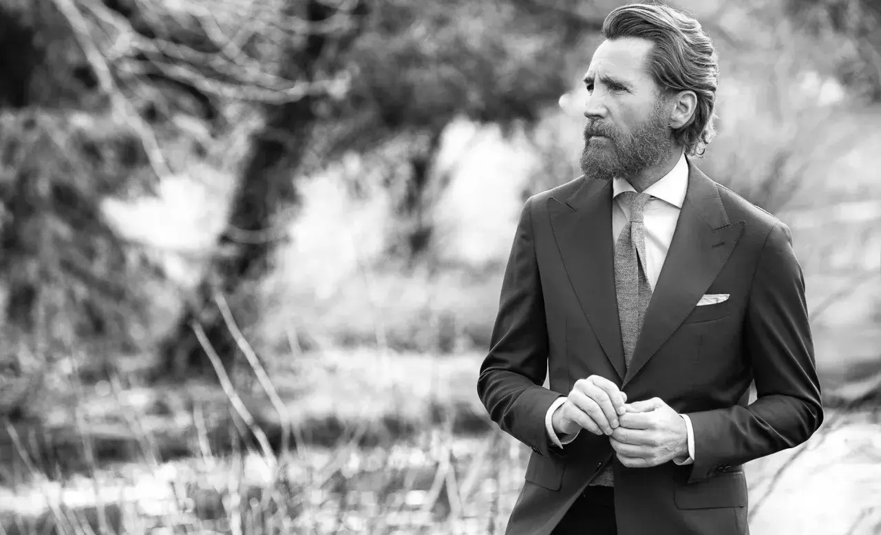Un homme barbu en costume sombre, chemise blanche et cravate grise, ajuste sa veste. Sa tenue avec short blanc homme est élégante.