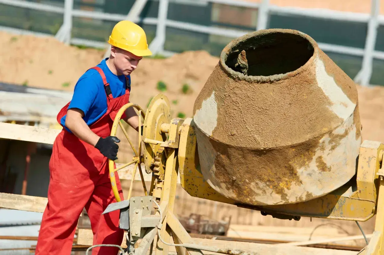 Robotnik w żółtym kasku i czerwonym ogrodniczkach obsługuje betoniarkę. Kluczowe są tu beton bez żwiru proporcje.