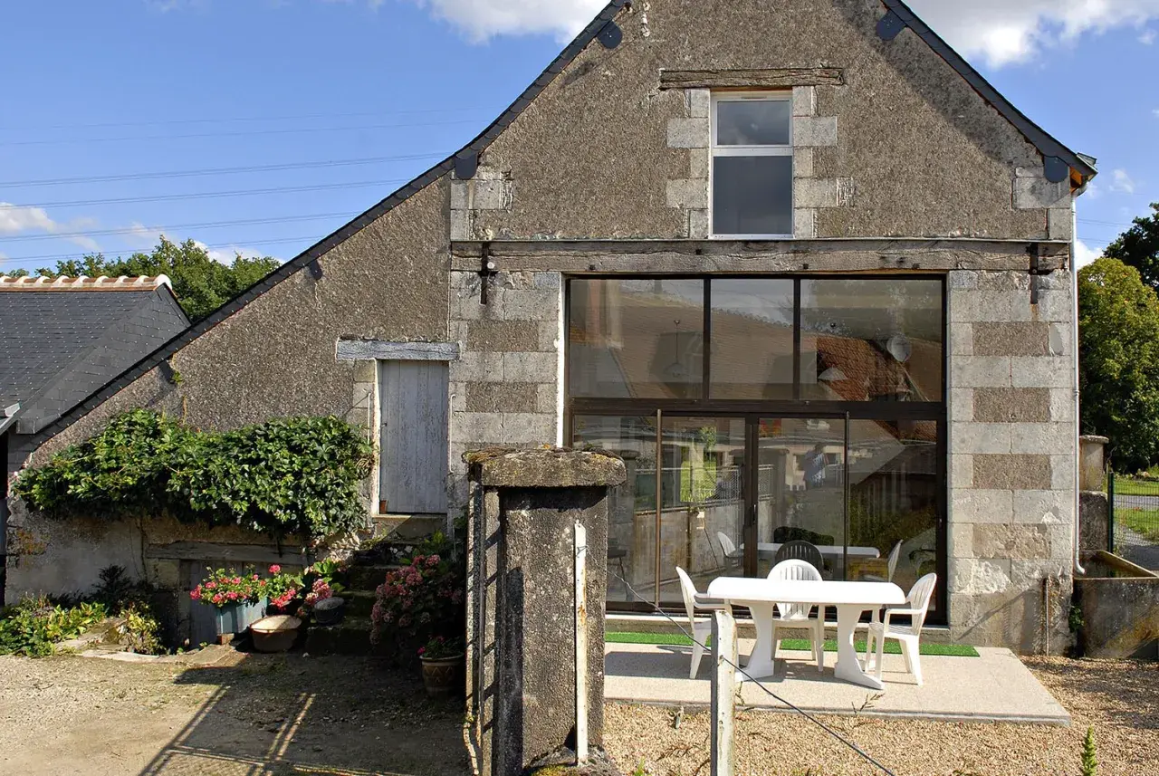 Ancienne grange en cours de rénovation, avec une façade en pierre et un grand mur vitré moderne. Une table et des chaises blanches sont installées sur la terrasse.