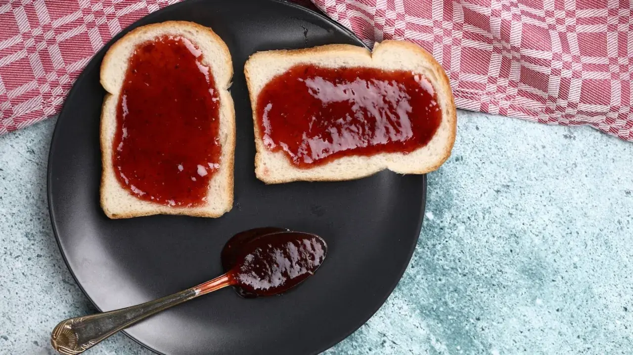 Zwei Scheiben Toast mit leckerer Jostabeeremarmelade und ein Löffel voll davon auf einem schwarzen Teller.