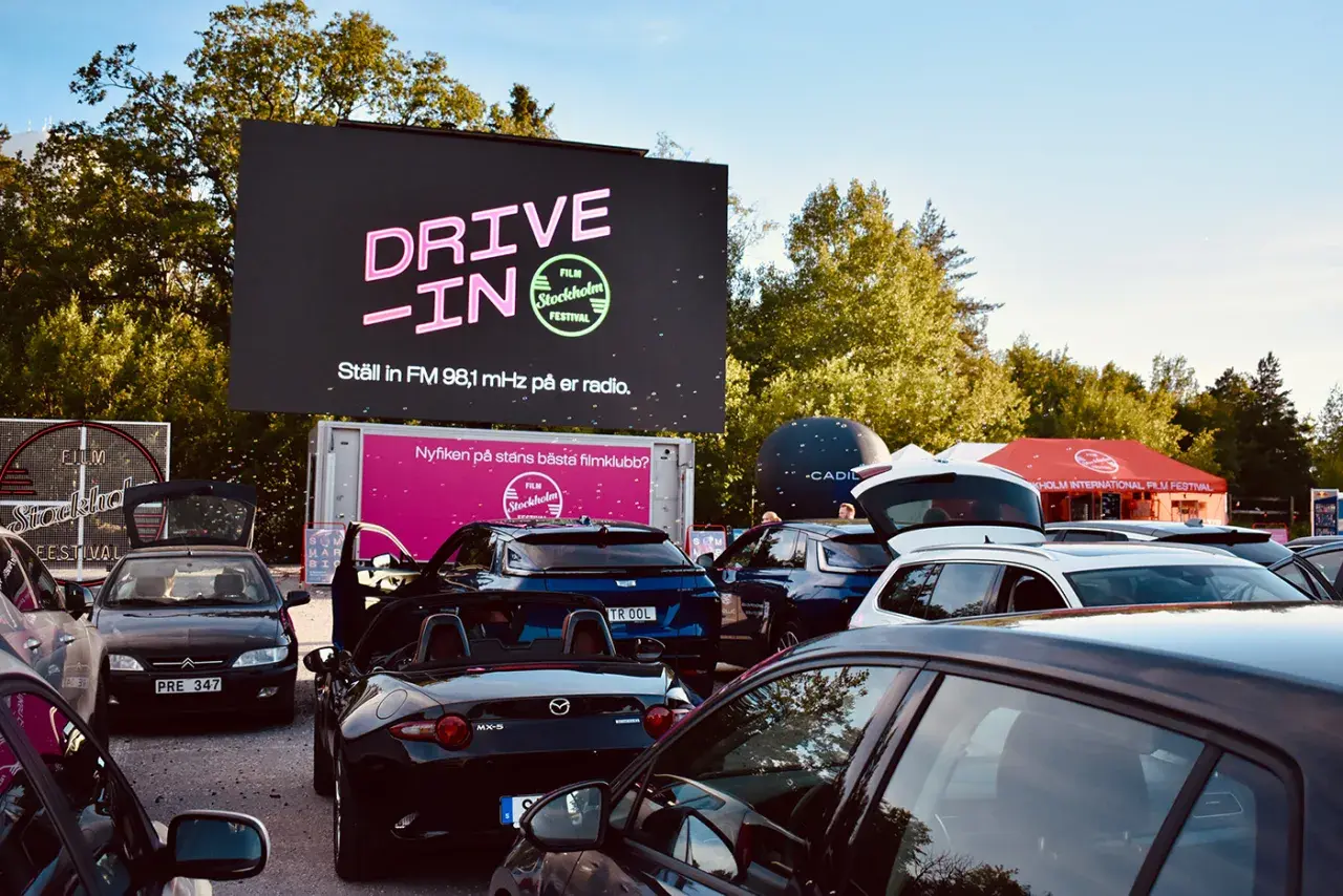 Bilar samlade vid en solvalla drive in bio. Stor skärm visar "DRIVE-IN FILM Stockholm Festival".