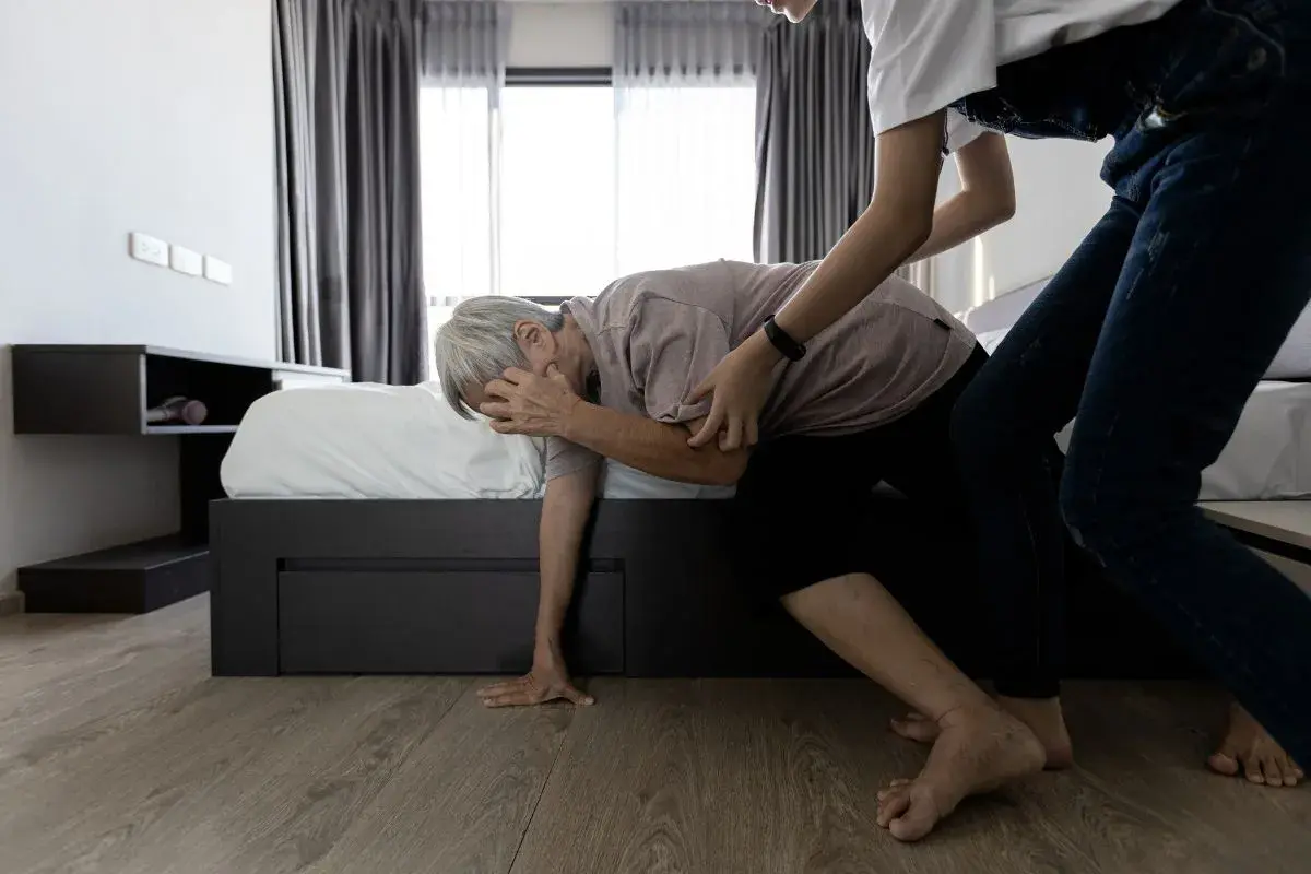 Assistenza a un anziano che accusa un cedimento delle gambe, con una persona che lo sorregge vicino al letto.