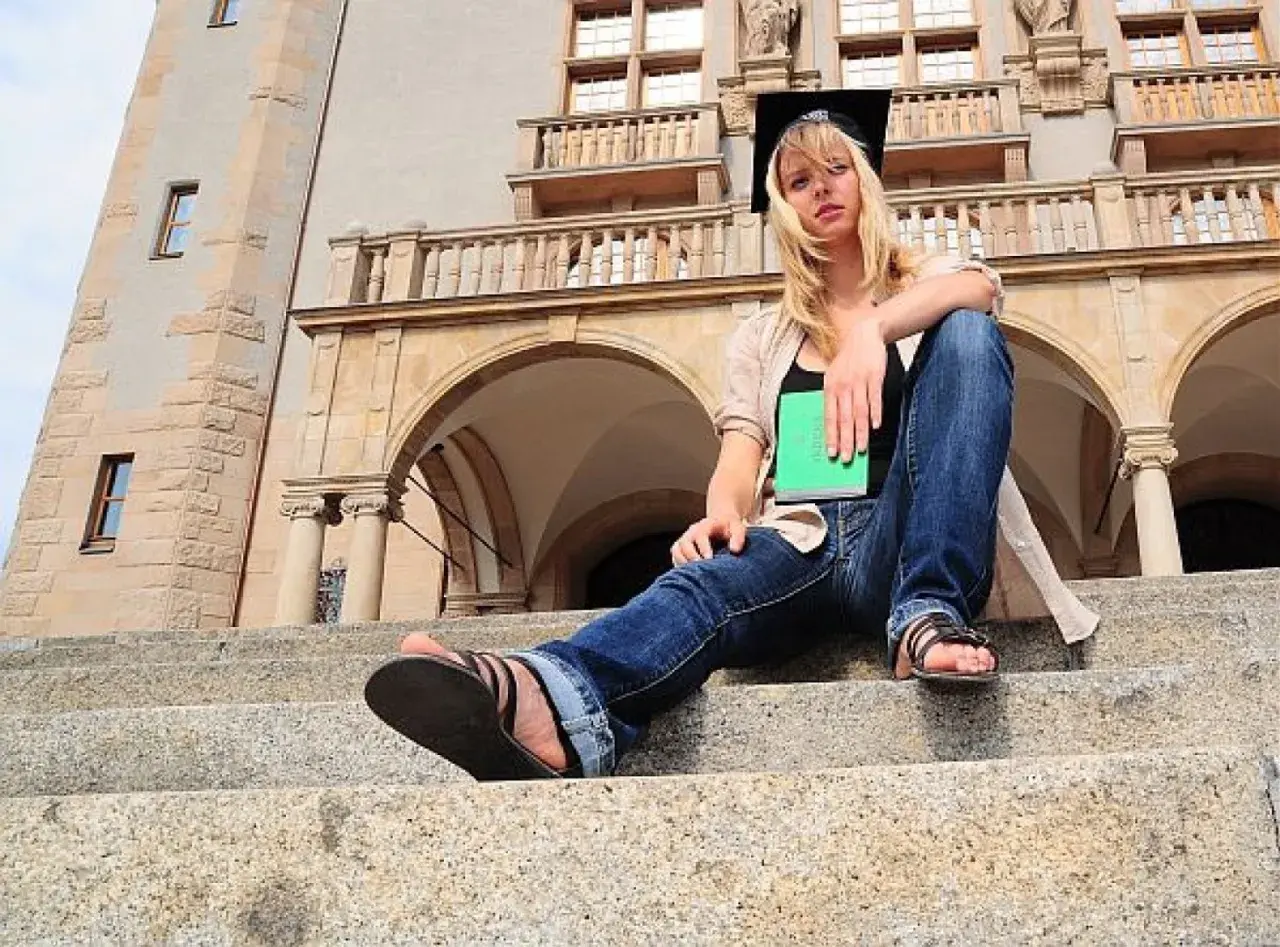Studentka w czepku absolwenta z zieloną książką, siedząca na schodach przed budynkiem uniwersyteckim. Marzy o studiach zaocznych w Poznaniu.