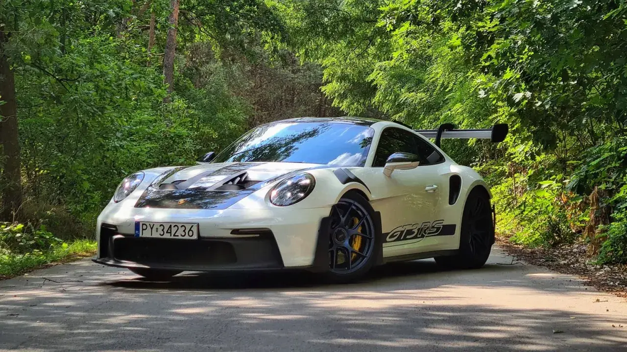 Biały Porsche 911 GT3 RS z czarnymi pasami na masce i bokach, zaparkowany na leśnej drodze. Jego cena jest wysoka, ale wrażenia z jazdy bezcenne.