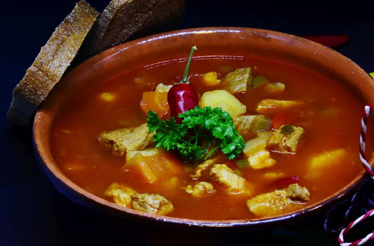 Gęsty bogracz wieprzowy z kawałkami mięsa, ziemniaków i papryki, ozdobiony papryczką chili i natką pietruszki. Obok kromki chleba.