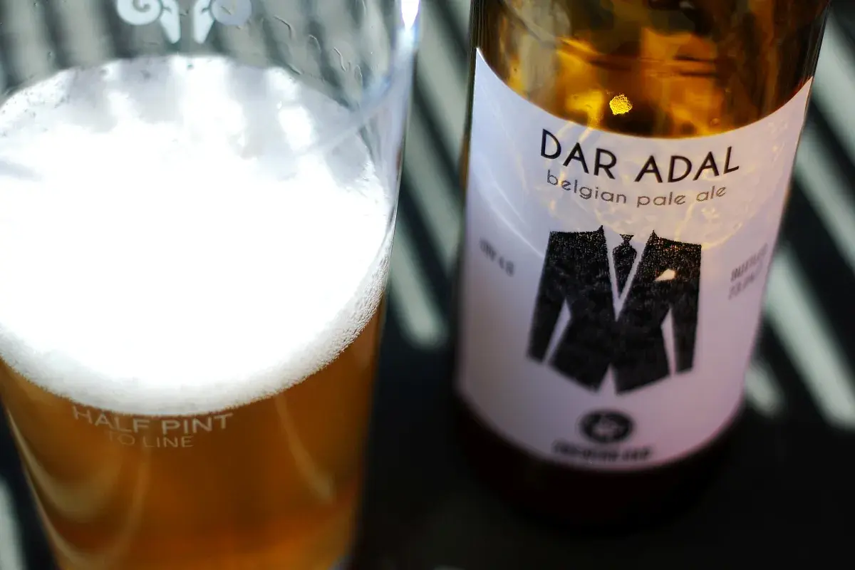 Bicchiere di birra dorata con schiuma bianca e bottiglia di Dar Adal belgian pale ale con etichetta a forma di giacca.