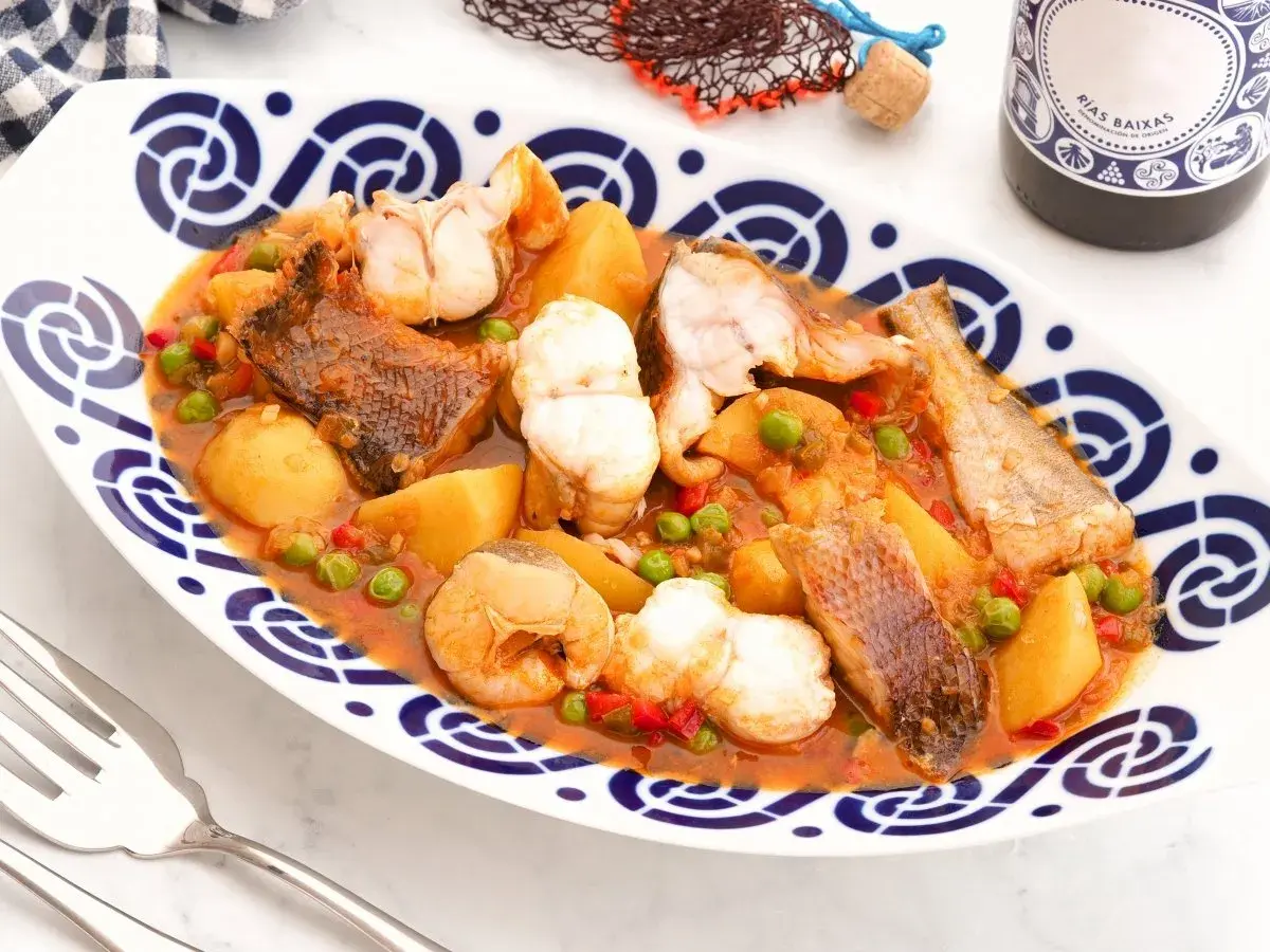 Caldeirada gallega servida en plato blanco con borde azul. Contiene trozos de pescado, patatas, guisantes y pimientos en una salsa rojiza.