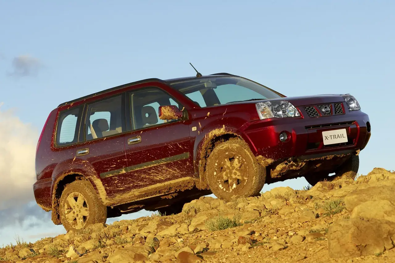 Czerwony Nissan X-Trail ubrudzony błotem podjeżdża pod skaliste wzniesienie.