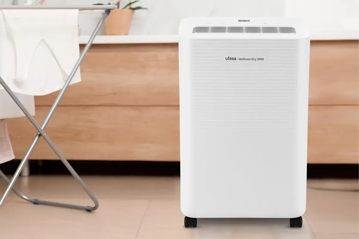 Un deshumidificador Ufesa Wellness Dry 2000, blanco y con ruedas, junto a un tendedero con ropa.