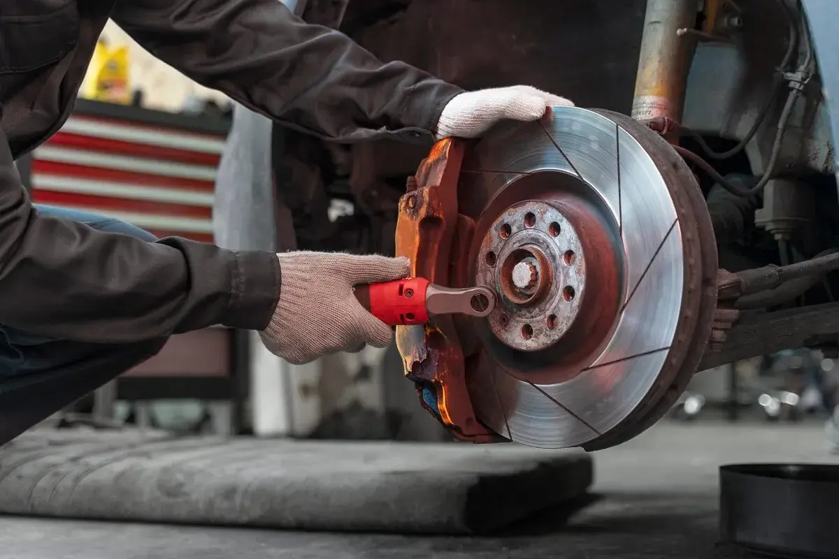 Mechanik w rękawiczkach dokonuje wymiany klocków hamulcowych, używając klucza do odkręcenia śruby zacisku.