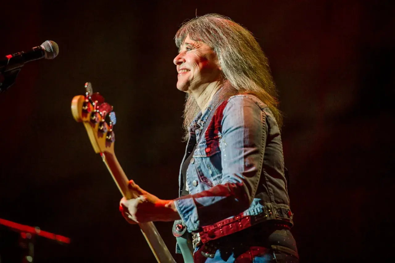 Suzi Quatro obecnie, z uśmiechem na twarzy, gra na gitarze basowej w dżinsowej kurtce.
