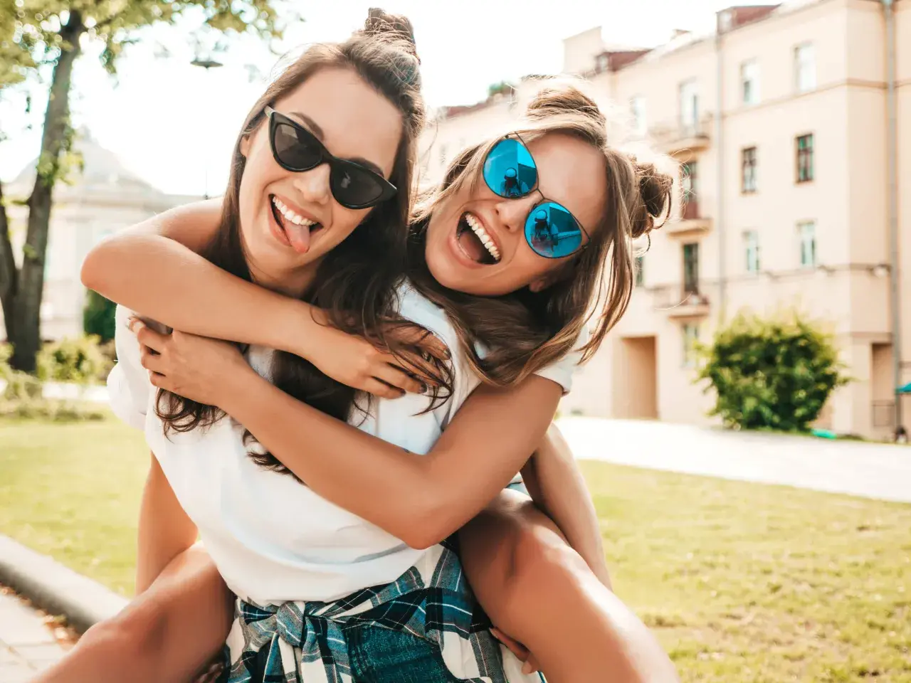 Dos amigas riendo y abrazándose, como si estuvieran escribiendo una carta a una amiga. Llevan gafas de sol y están al aire libre.