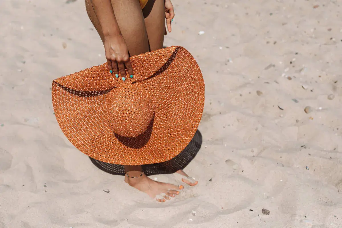 Donna con cappello di paglia arancione sulla sabbia, un'idea per come mantenere l'abbronzatura.