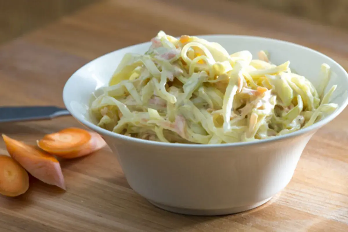 En skål med krämig, godaste coleslaw med strimlad vitkål och morötter. Bredvid ligger några morotsskivor.