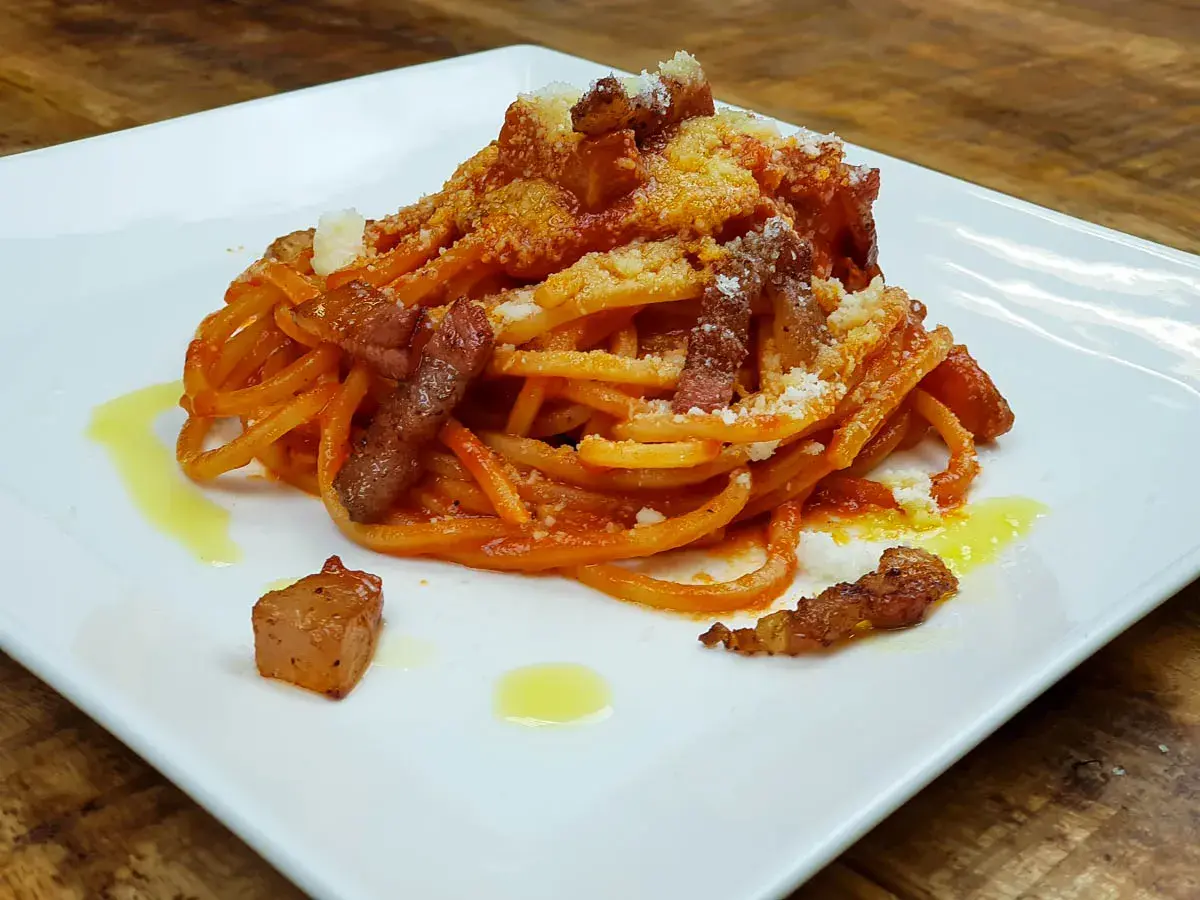Bucatini con salsa amatriciana, cubos de guanciale crujiente y queso rallado, servido en plato blanco.
