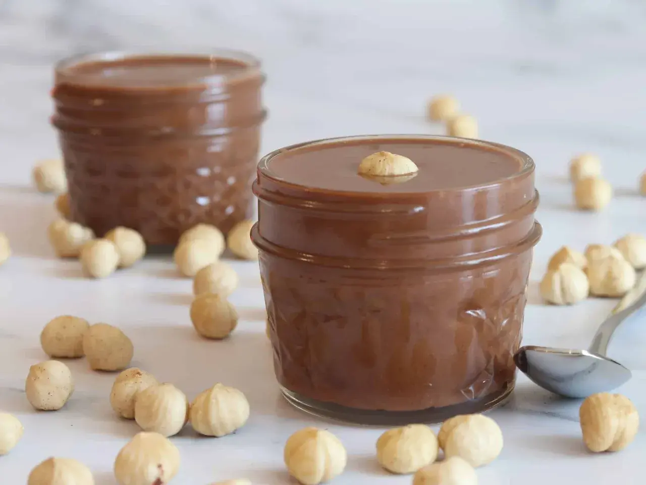 Dos tarros de deliciosa nutella vegana rodeados de avellanas. Una cucharilla plateada descansa cerca.