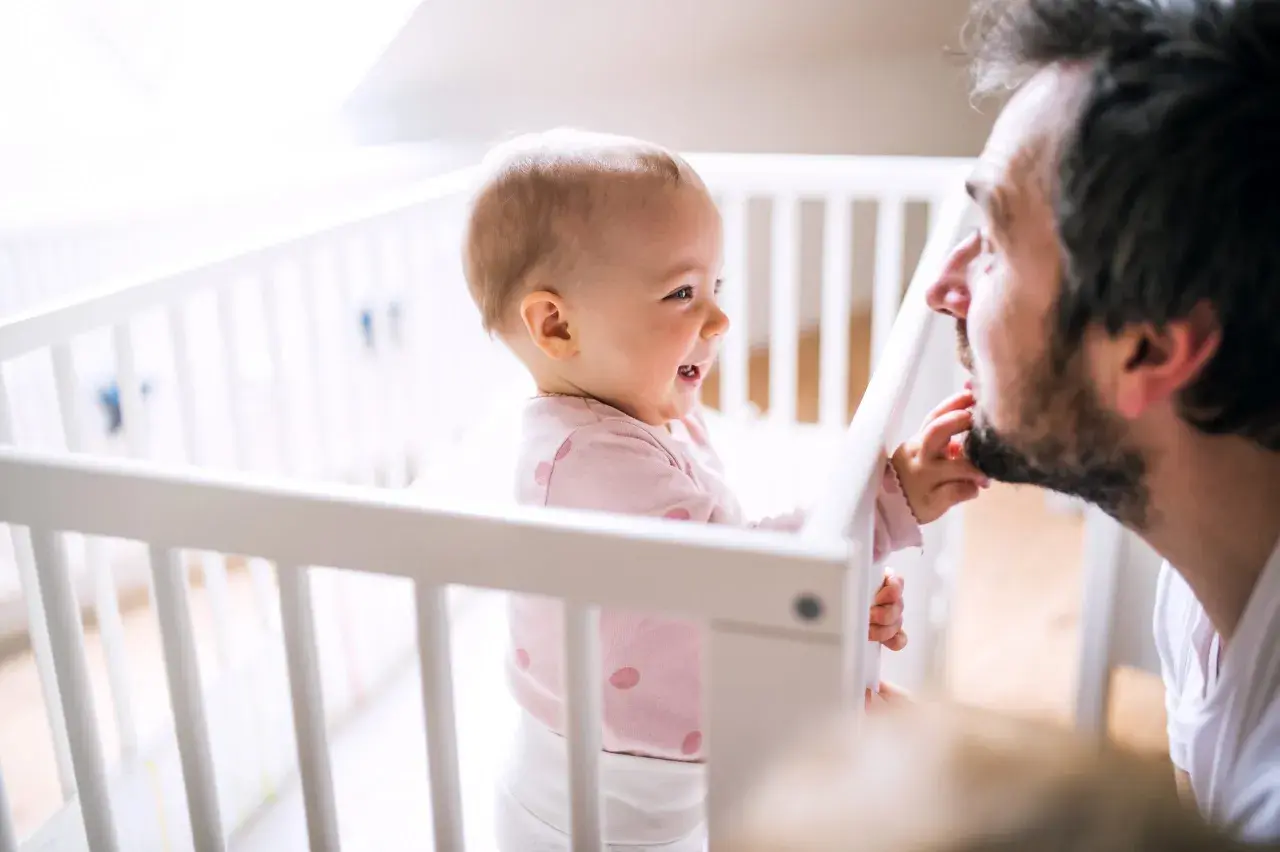 Een lachende baby in een ledikantje reikt naar de baard van haar vader. Pure liefde tussen vader en dochter.
