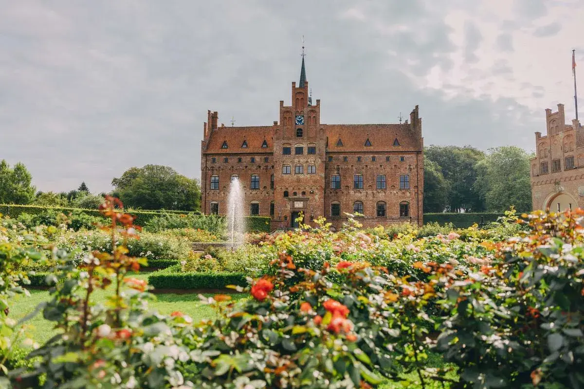 En vacker tegelbyggnad med torn och en fontän i en blomstrande trädgård. En dröm för en vecka på Fyn.