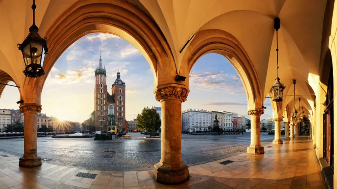 Krakau Geheimtipps: Blick durch Arkaden auf den Marktplatz mit Marienkirche im Sonnenaufgang. Ein malerischer Ort für Entdeckungen.