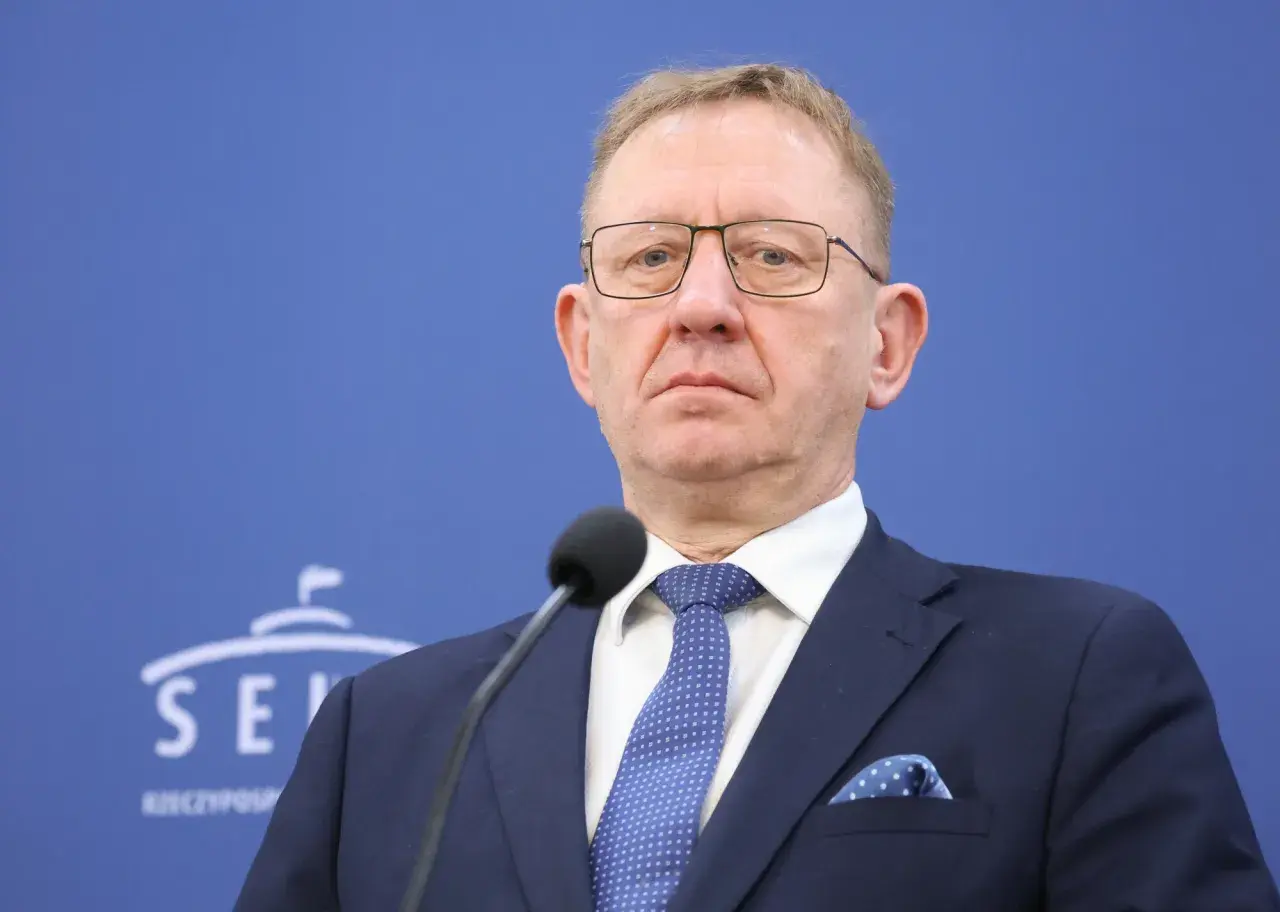 Mężczyzna w okularach i granatowym garniturze przed mikrofonem na tle niebieskiej ściany z logo Senatu.