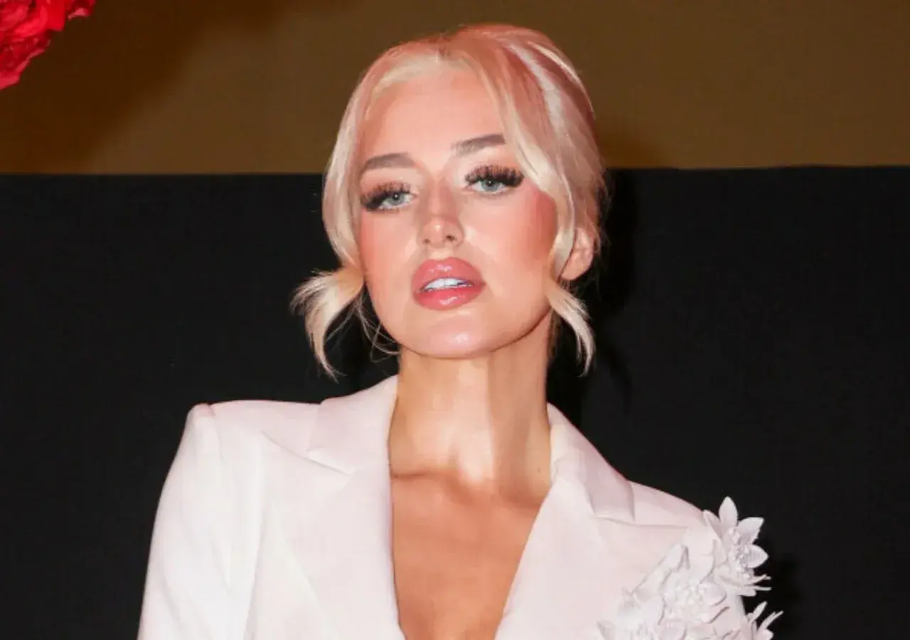 Caroline Derpienski w białej marynarce, z blond włosami upiętymi w dwa kucyki. Jej makijaż podkreśla usta i oczy.