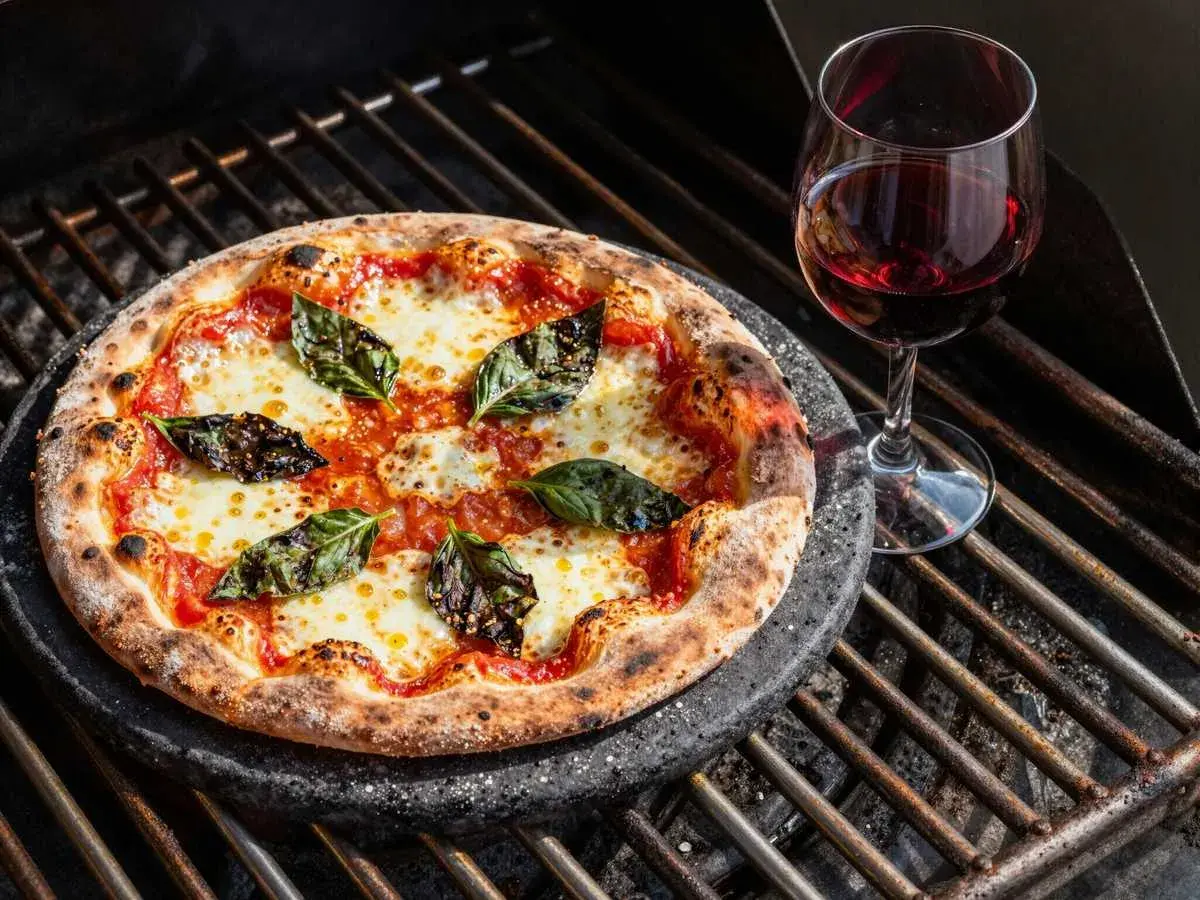 Pizza mit langer Gehzeit auf dem Grill, belegt mit Tomaten, Mozzarella und Basilikum, daneben ein Glas Rotwein.