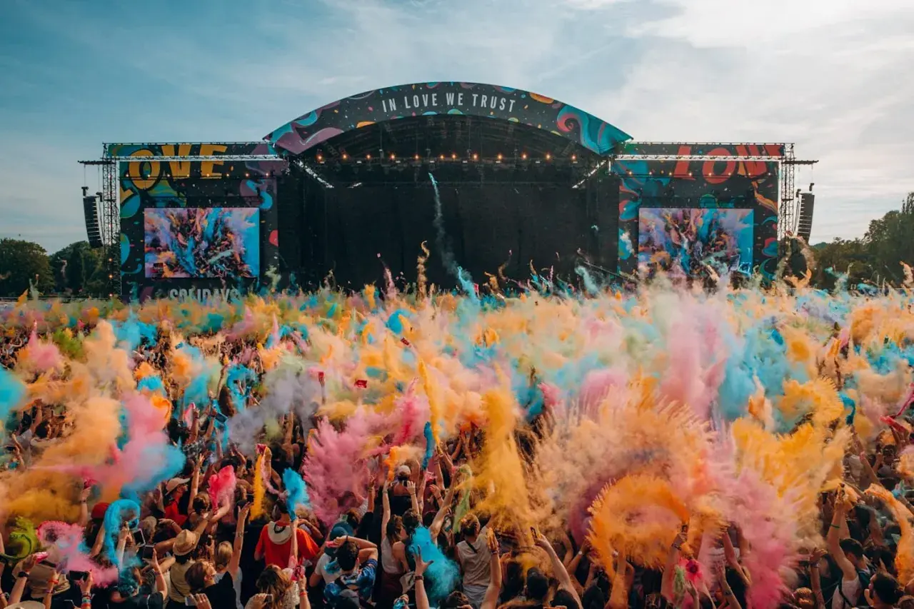 Une foule en liesse lance de la poudre colorée pour créer un festival de musique inoubliable. L'ambiance est électrique sous la scène "IN LOVE WE TRUST".