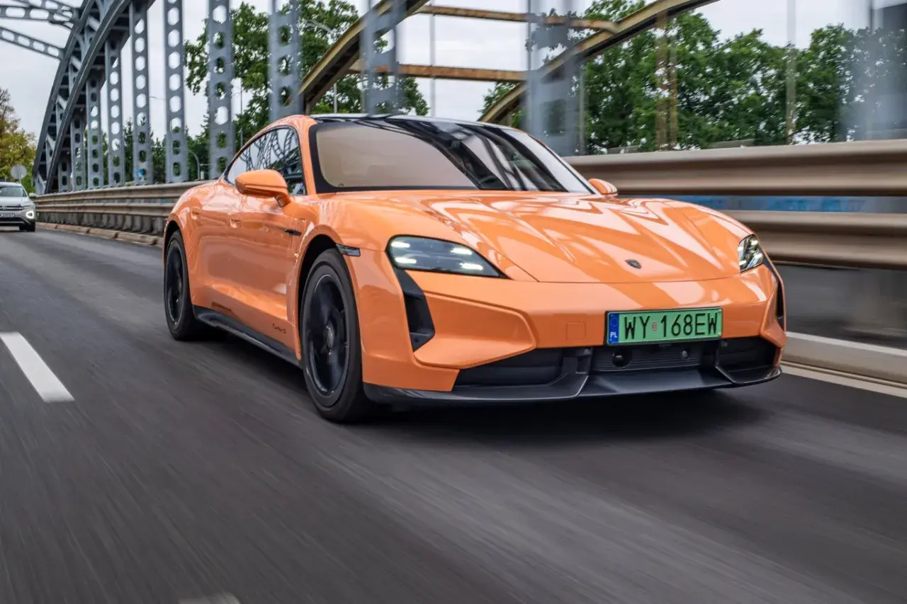 Pomarańczowy Porsche Taycan Turbo S pędzi po moście, jego sportowy design podkreślony przez dynamiczne linie.
