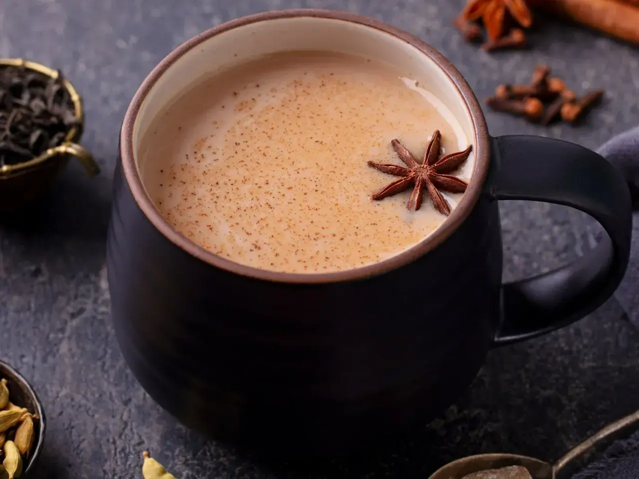 Aromaticzny napój, idealny na chłodne dni. Gorący **masala chai** w czarnej filiżance, ozdobiony anyżem. Obok liście herbaty, kardamon i goździki.