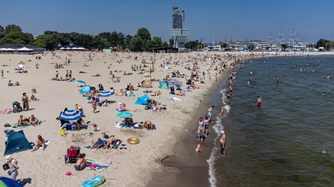 Słoneczna plaża w jednej z miejscowości koło Gdyni, pełna wypoczywających ludzi. W tle widać drapacz chmur i żaglowce.