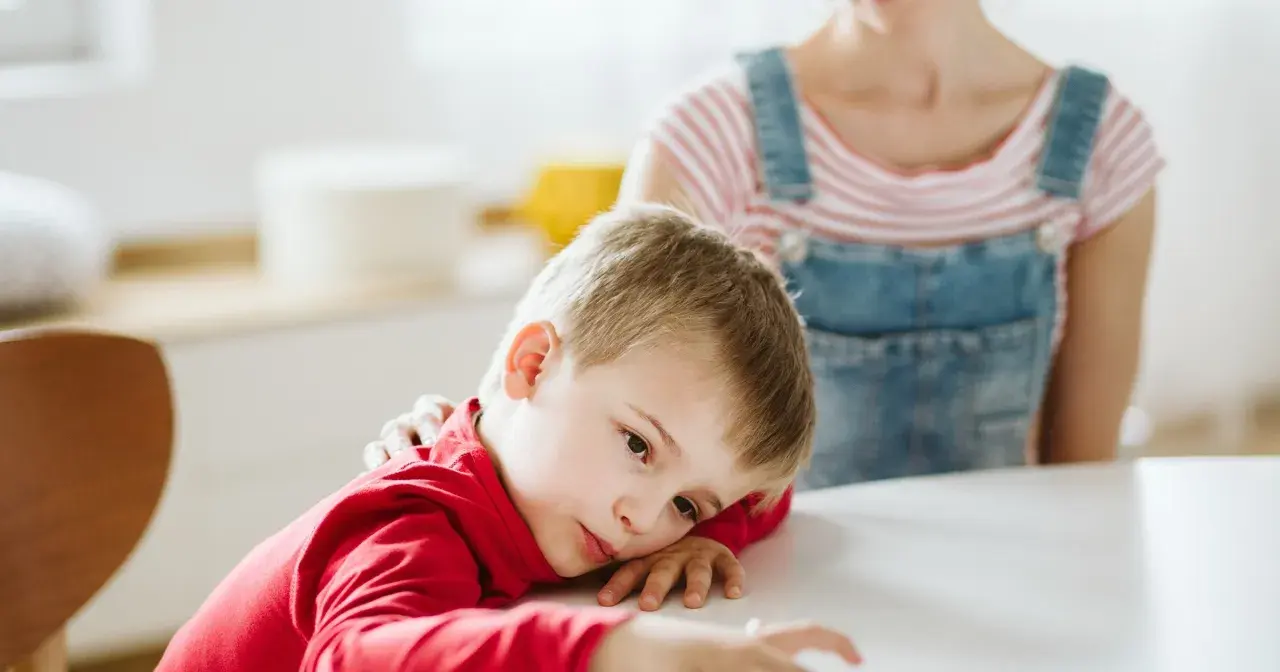 Smutny chłopiec z ADHD opiera głowę na stole, podczas gdy matka kładzie mu rękę na ramieniu.