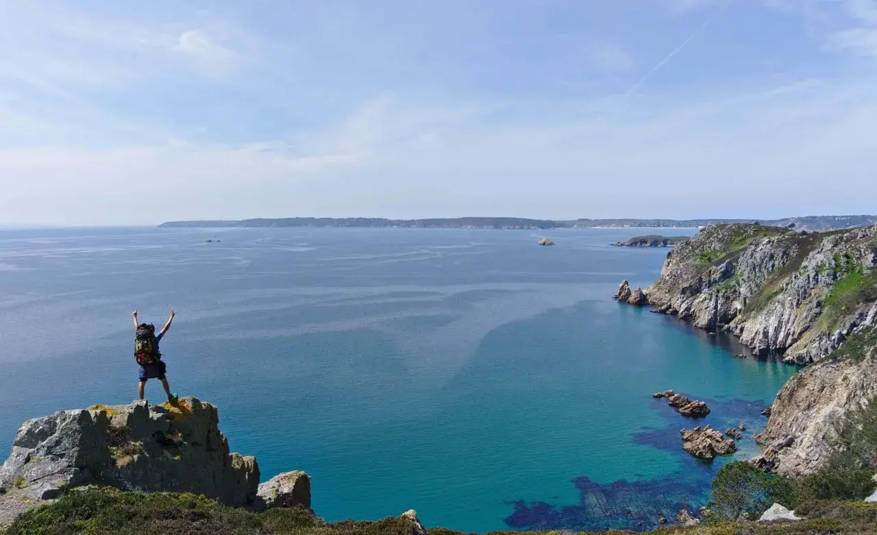 Randonneur victorieux au sommet d'un rocher, bras levés, dominant la mer turquoise et les falaises de GR34 Crozon.