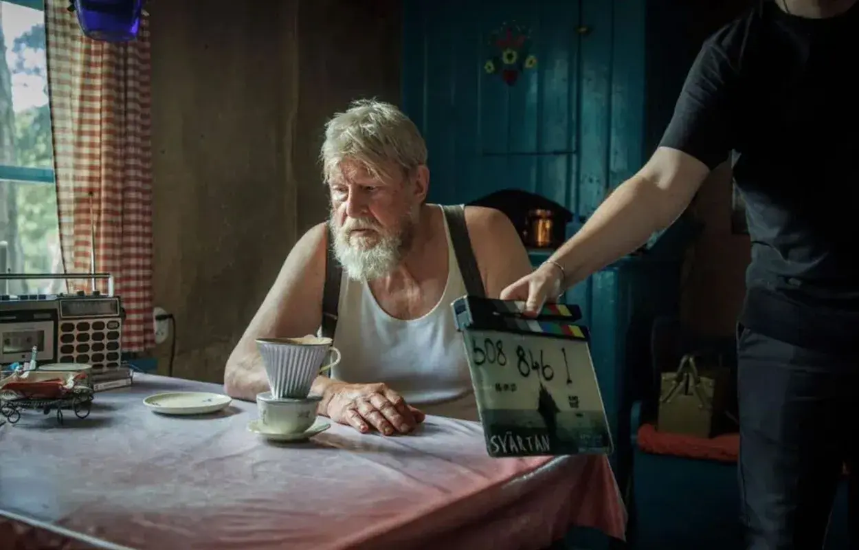 Rolf Lassgård sitter vid ett bord med en kaffebryggare. En person håller upp en filmklappa för en ny serie med Rolf Lassgård.
