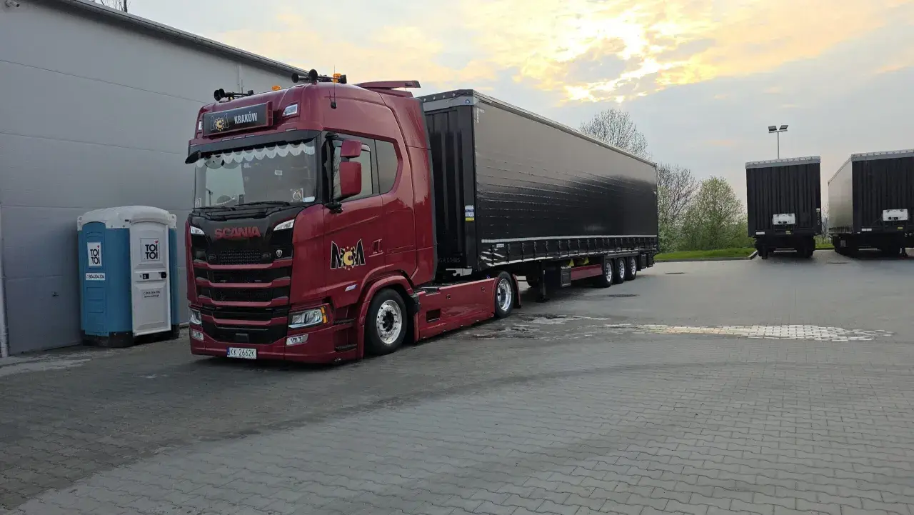 Czerwona Scania z naczepą gotowa do przewozu drobnicowego. Obok stoi niebieska toaleta przenośna.