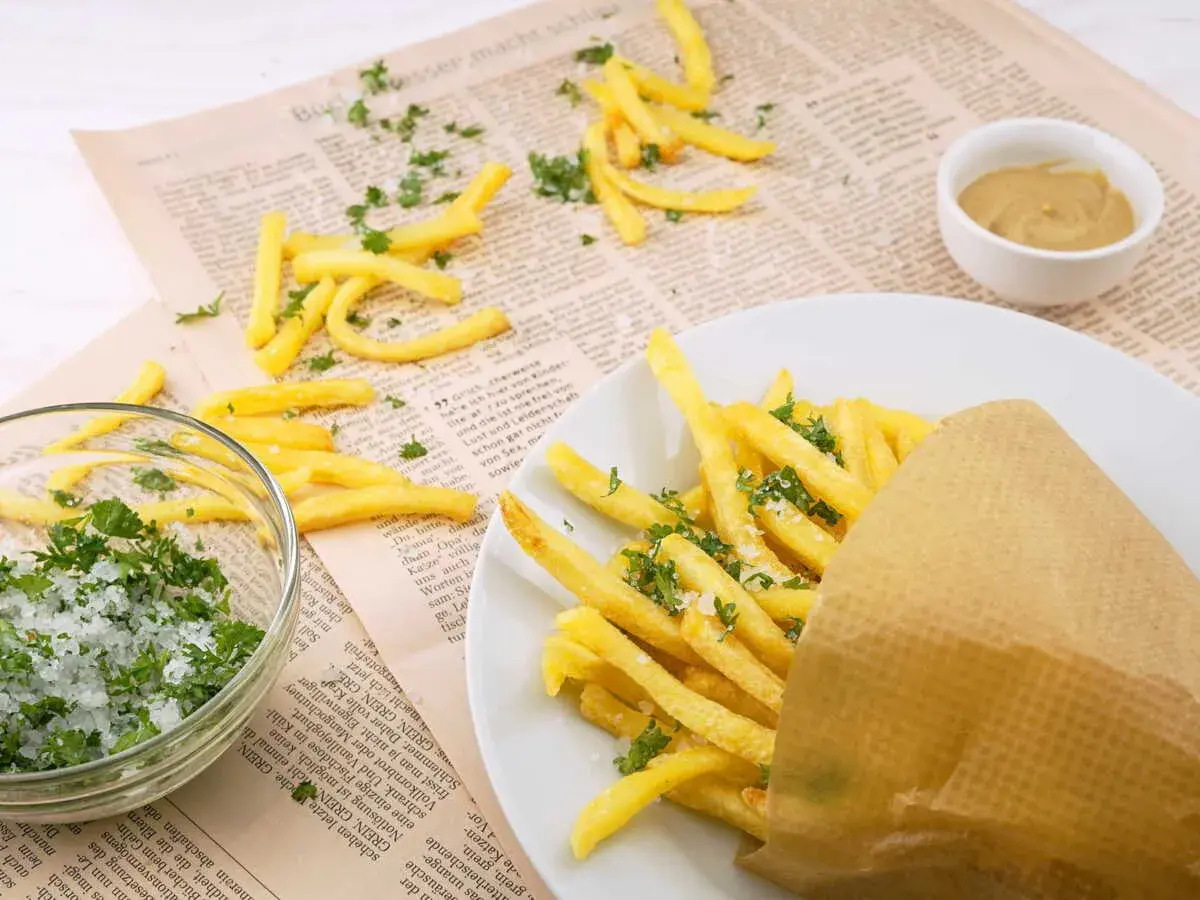 Des frites dorées, saupoudrées de persil et de sel, servies dans un cornet en papier. À côté, un bol de sel et de persil, et une petite coupelle de moutarde. Un délice qui rappelle les sardines frites.