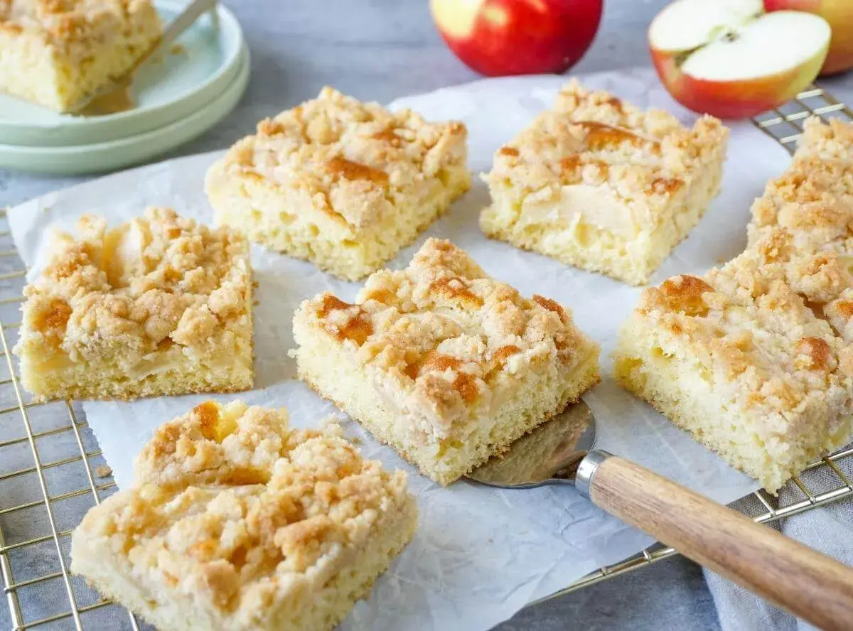 Saftiger Apfelkuchen vom Blech mit Streuseln, frisch gebacken und in Stücke geschnitten. Daneben liegen Äpfel.