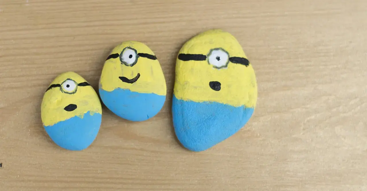 Trzy kamienie polne pomalowane na żółto i niebiesko, przypominające Minionki. Zabawne malowanie kamieni polnych.