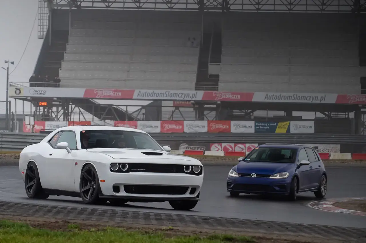 Biały Dodge Challenger i niebieski Volkswagen Golf na torze Słomczyn. W tle trybuny i banery reklamowe.