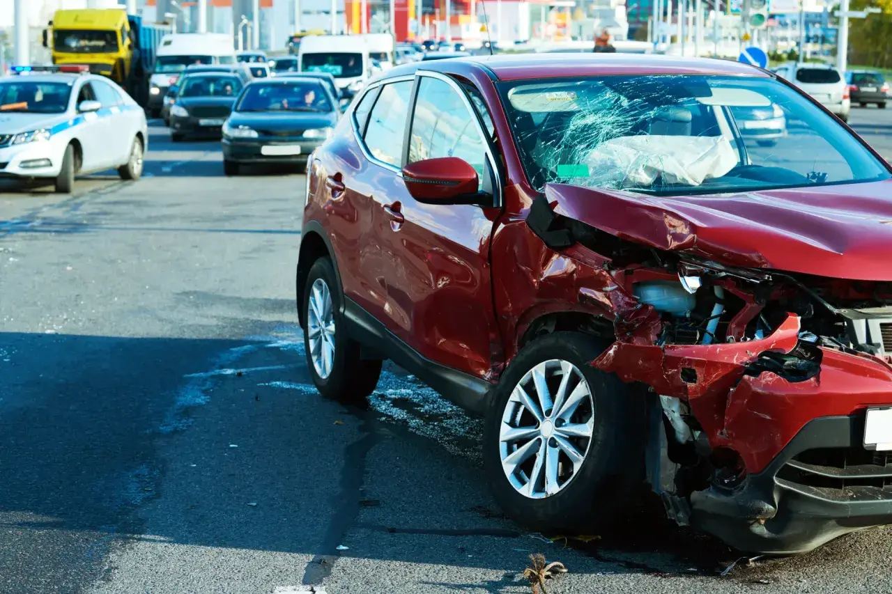 Wypadek drogowy. Czerwony samochód po kolizji, zniszczony przód i pęknięta szyba. Na drodze inne pojazdy, w tym radiowóz. Może to być sytuacja wymagająca **umowy na zastępstwo** kierowcy.