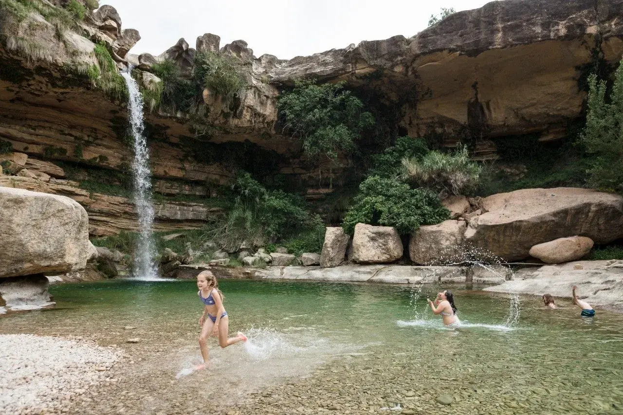 Niños disfrutan en una **piscina natural** bajo una cascada, rodeada de rocas y vegetación.