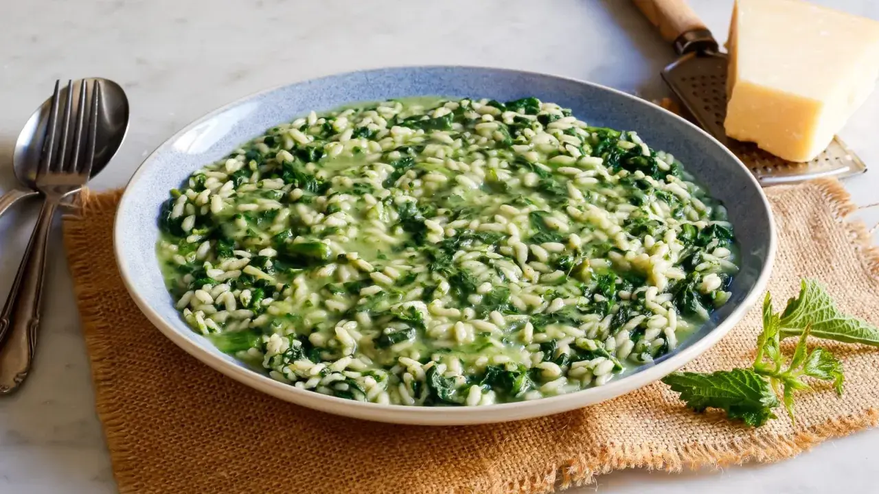 Un cremoso risotto con ortiche, servito in un piatto blu, con forchetta e coltello a lato. Un pezzo di parmigiano e una grattugia completano la scena.