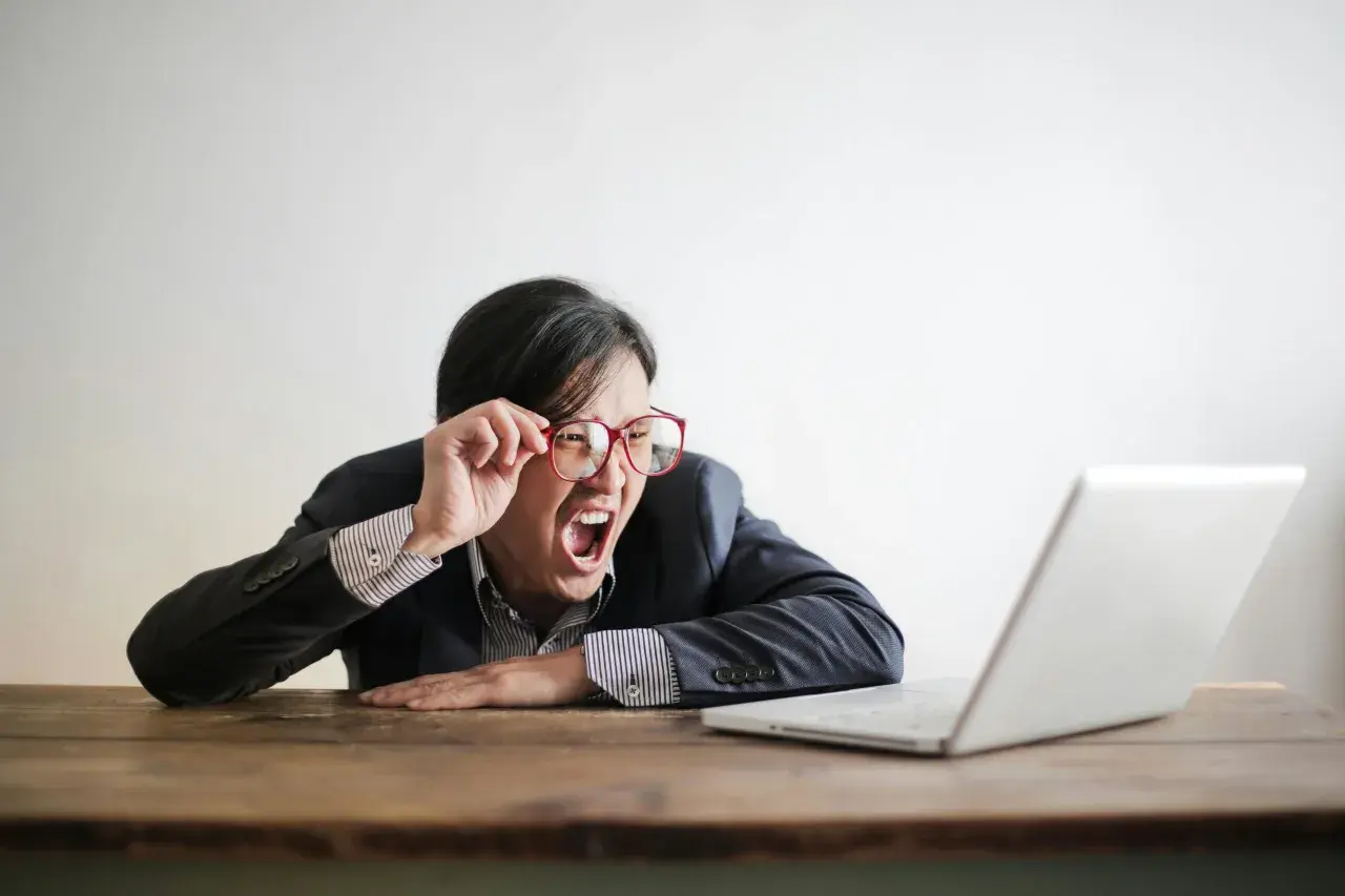 Mann mit Brille schreit frustriert vor Laptop. Vielleicht lernt er gerade, wie funktioniert ein Fernstudium, und ist überfordert.